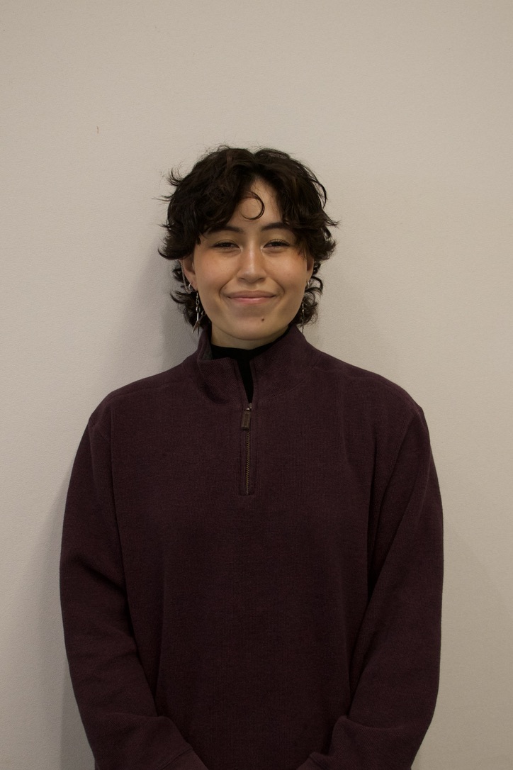 Person with short curly dark hair wearing a dark purple zip-up sweater, standing against a plain beige wall, smiling gently.