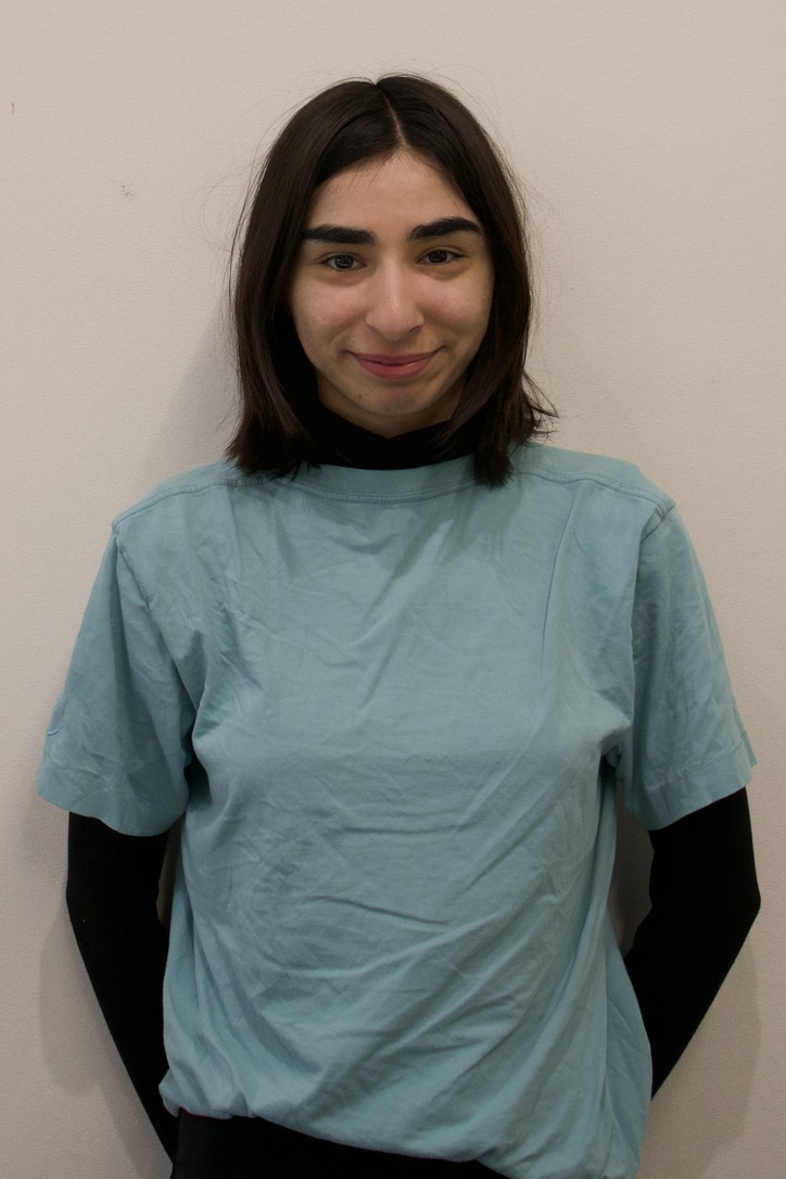 Young person with shoulder-length brown hair wearing a light blue t-shirt over a black long-sleeve shirt standing against a beige wall.