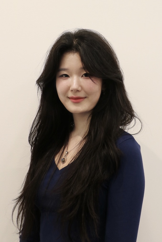 Smiling young woman with long dark hair wearing a navy blue top and a pendant necklace standing against a plain light background.