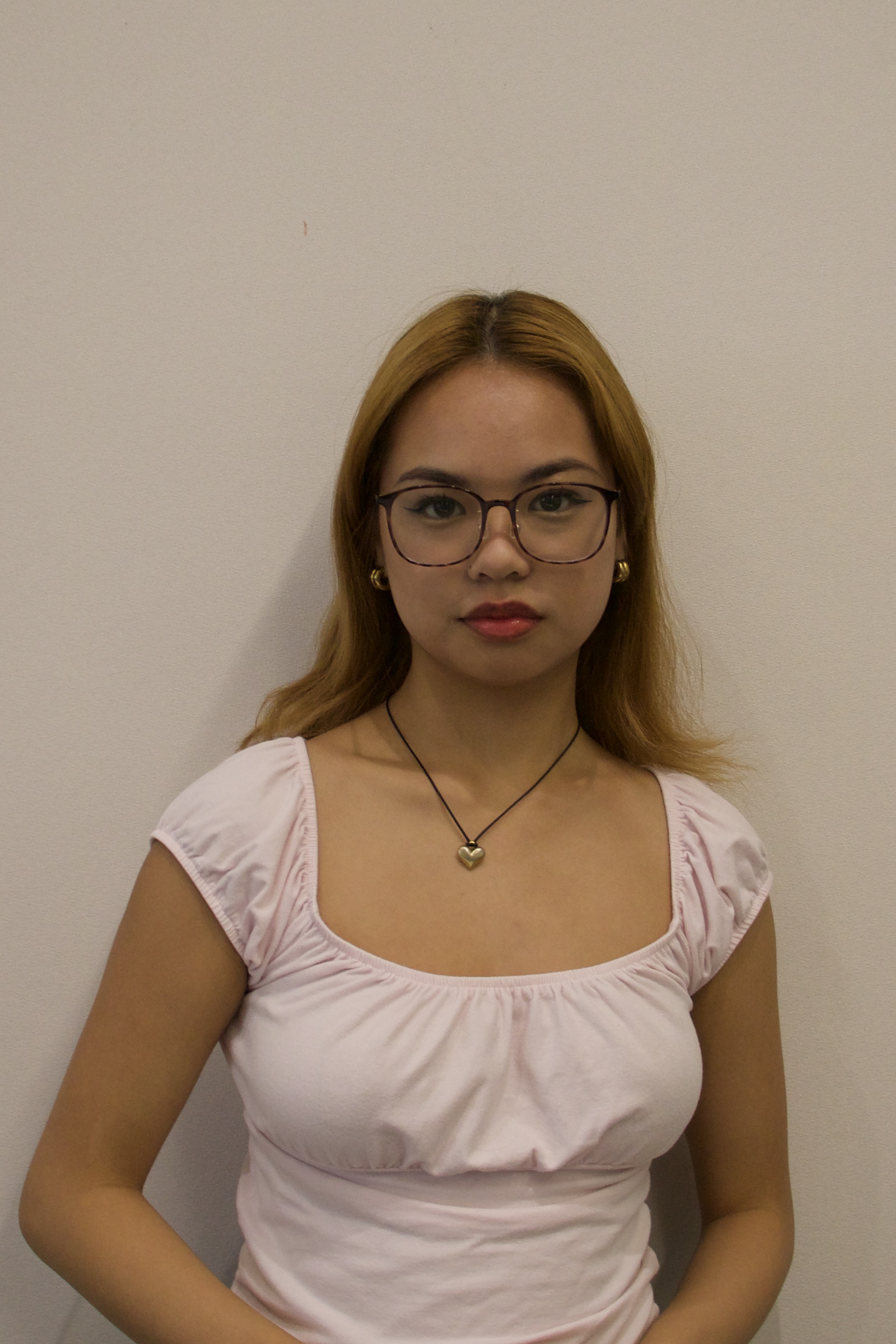 Young woman with long blonde hair, wearing glasses, gold hoop earrings, a heart pendant necklace, and a light pink top.