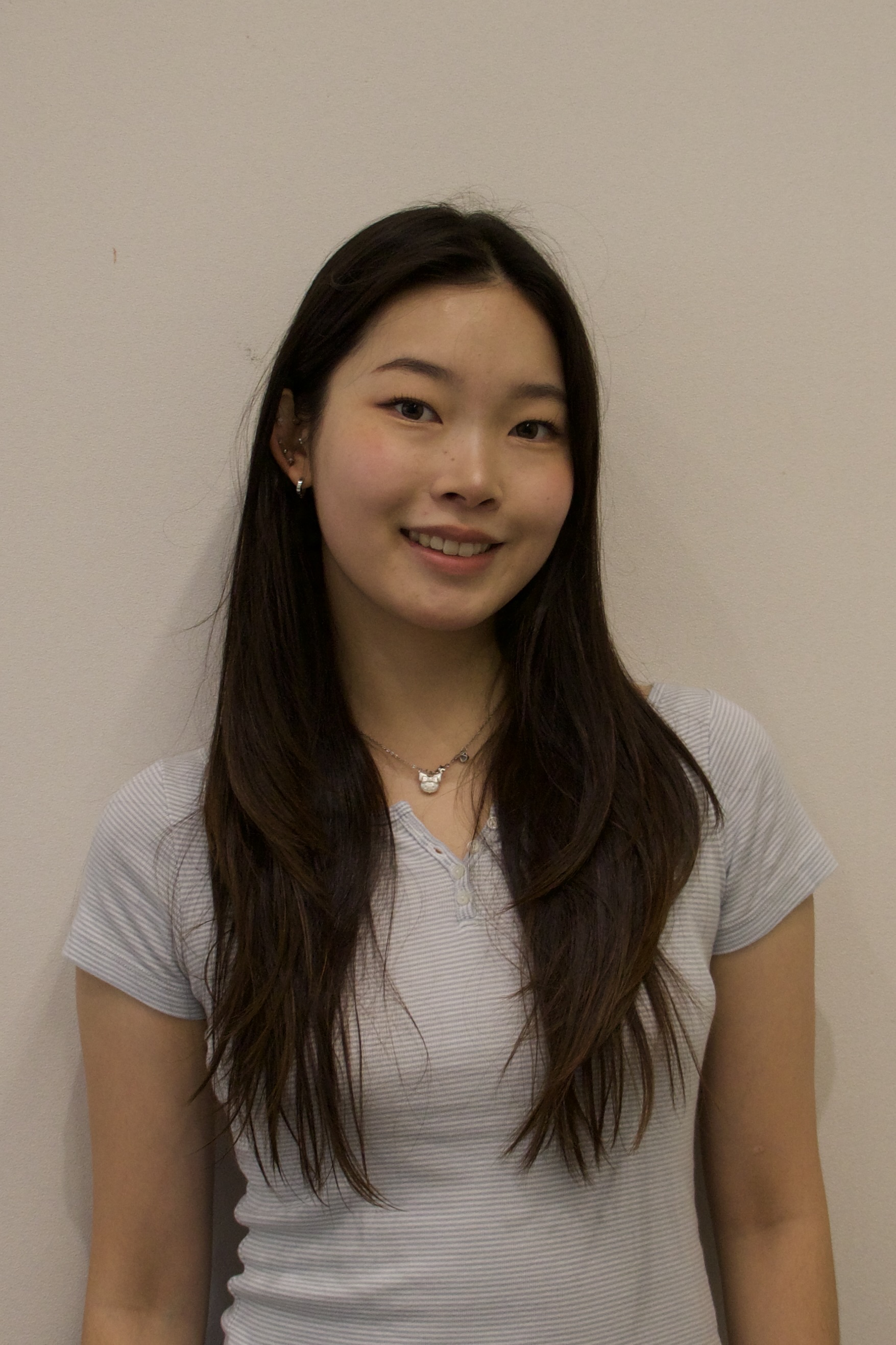 Young woman with long dark hair wearing a light gray striped shirt smiling in front of a plain beige wall.