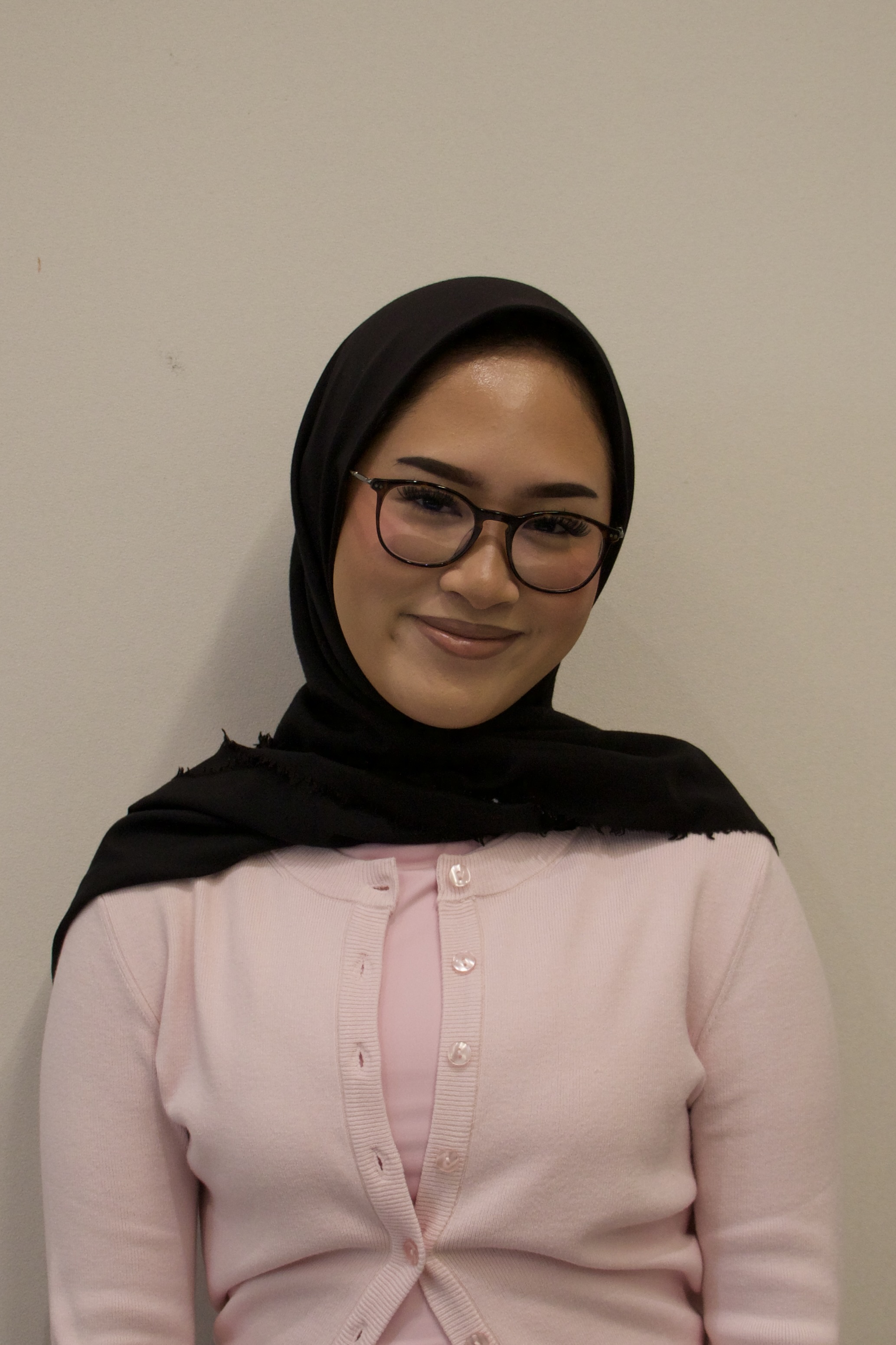 Smiling woman wearing a black hijab, glasses, and a light pink cardigan against a plain background.