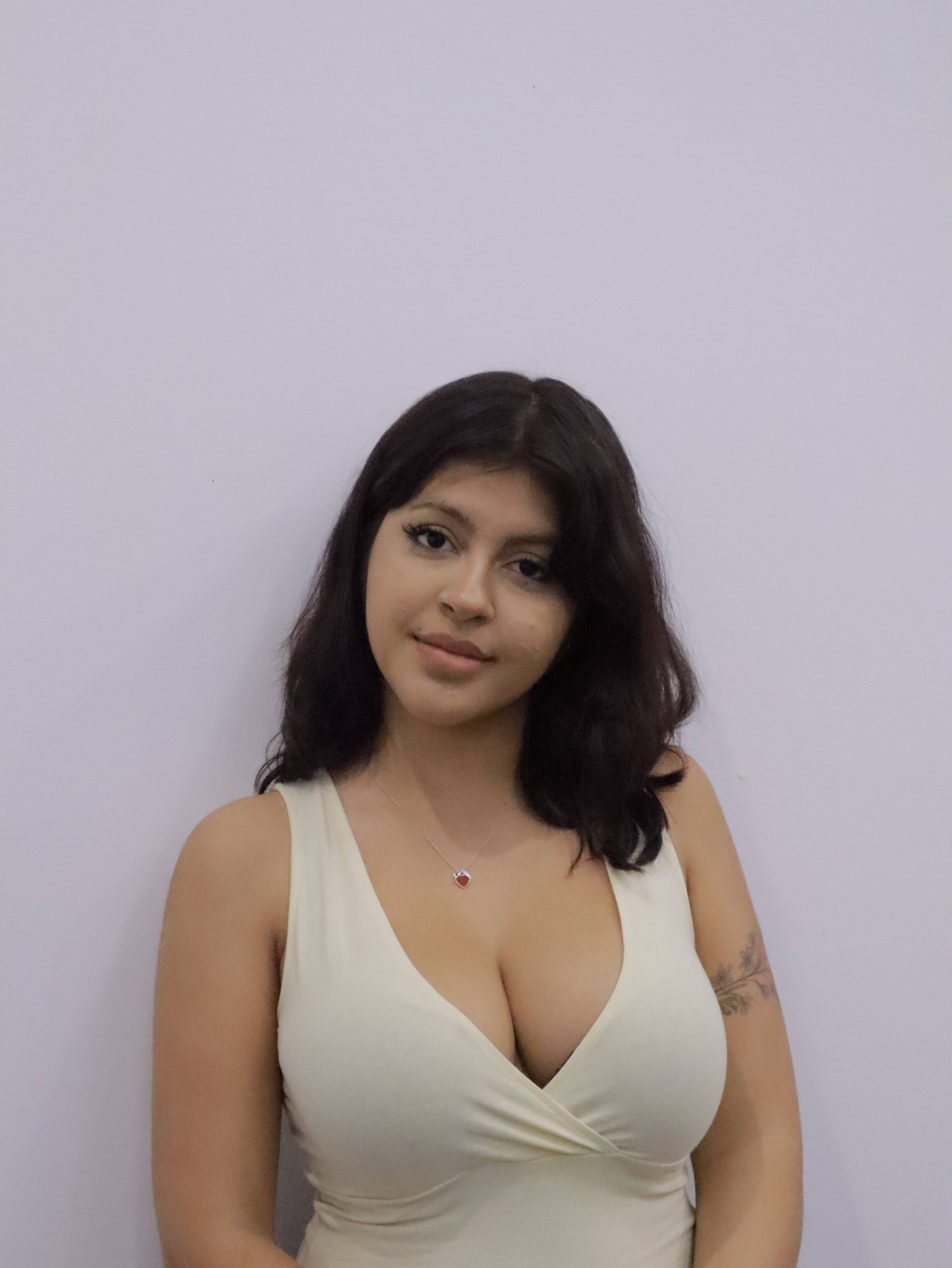Young woman with dark hair wearing a light-colored sleeveless top and a heart-shaped pendant necklace, standing against a plain light purple wall.