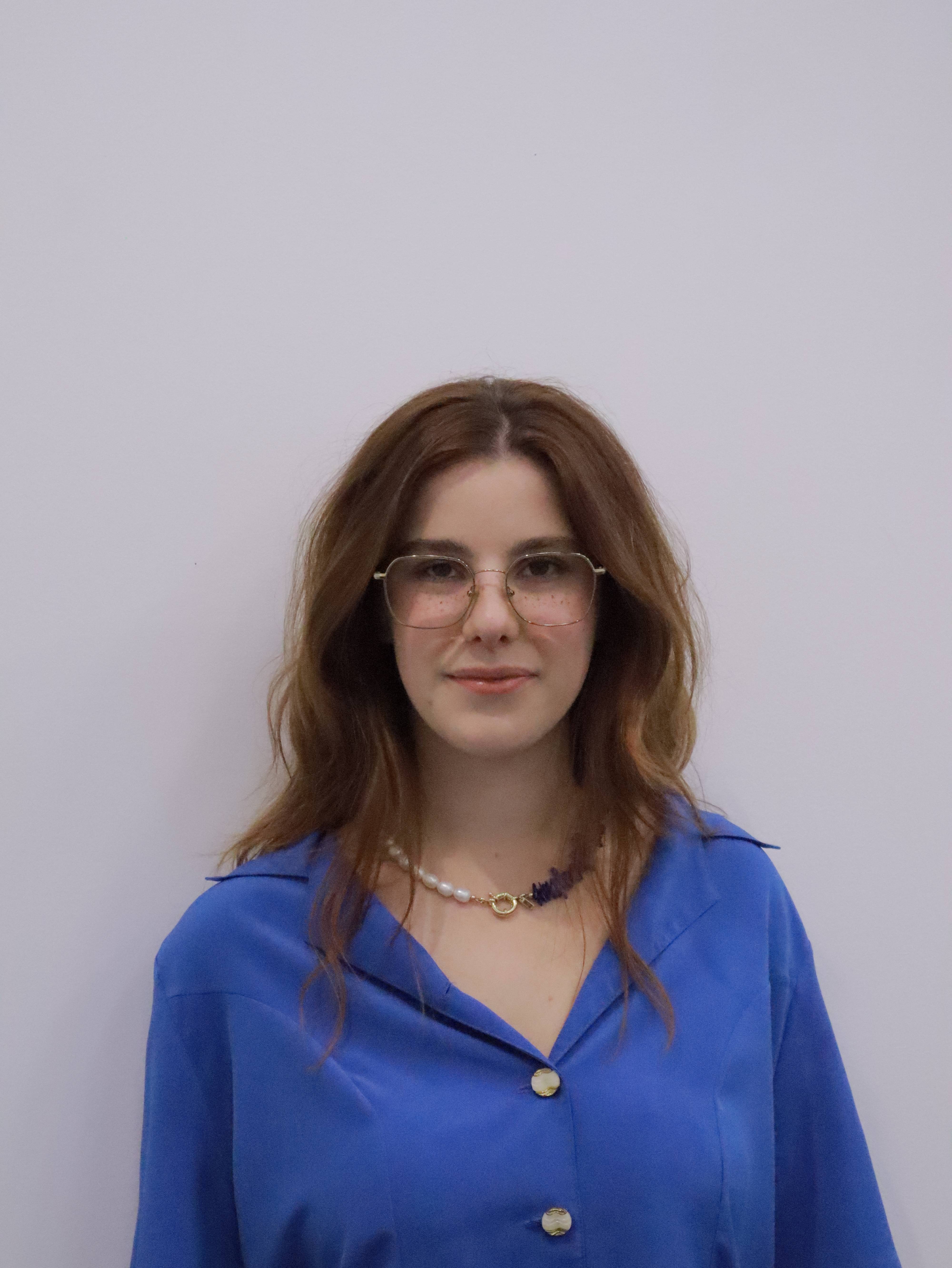 Young woman with shoulder-length brown hair, wearing glasses, a blue button-up shirt, and a pearl necklace against a plain light background.