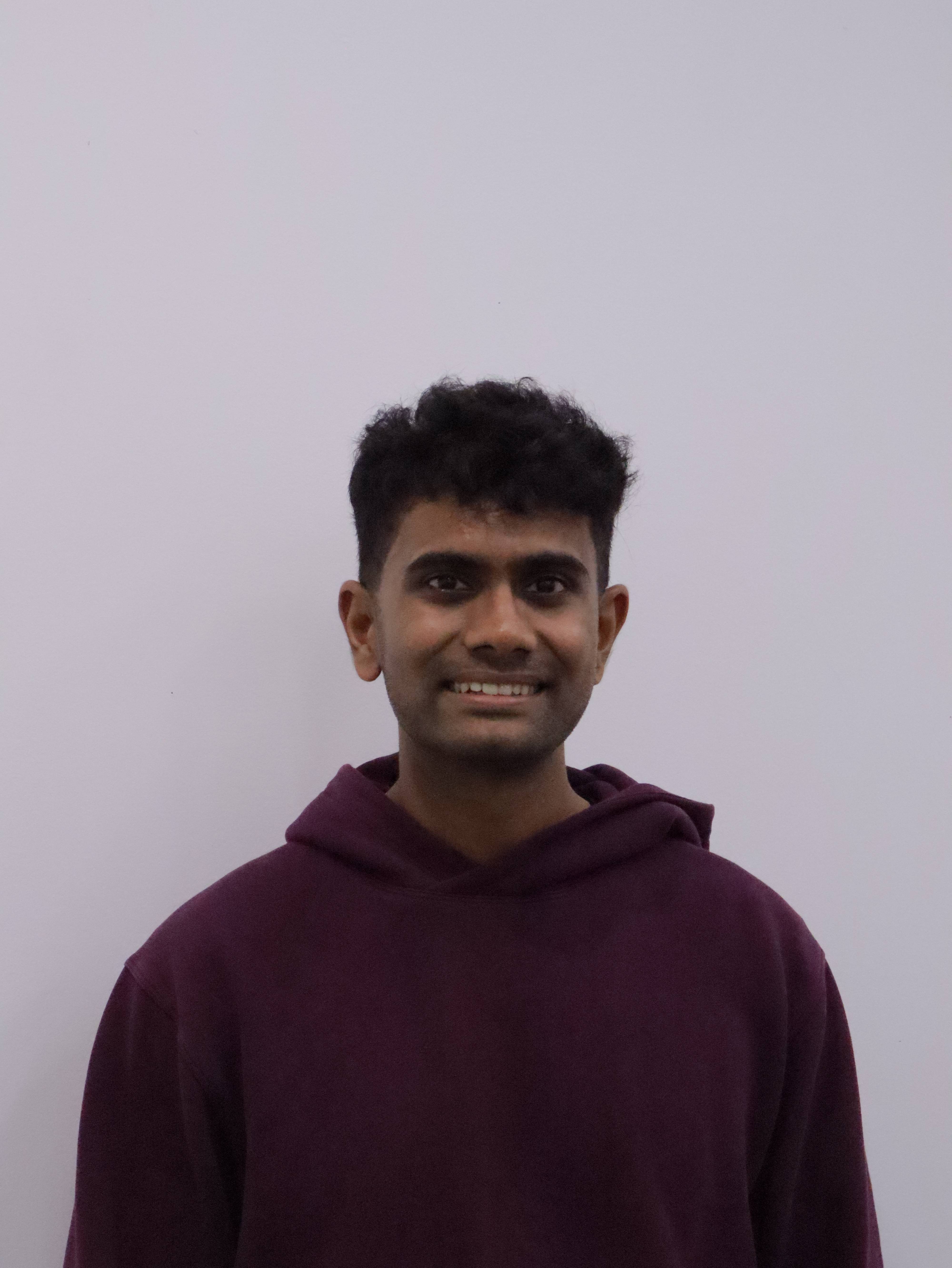 Portrait of a young man with curly black hair wearing a dark purple hoodie, smiling against a plain light gray background.