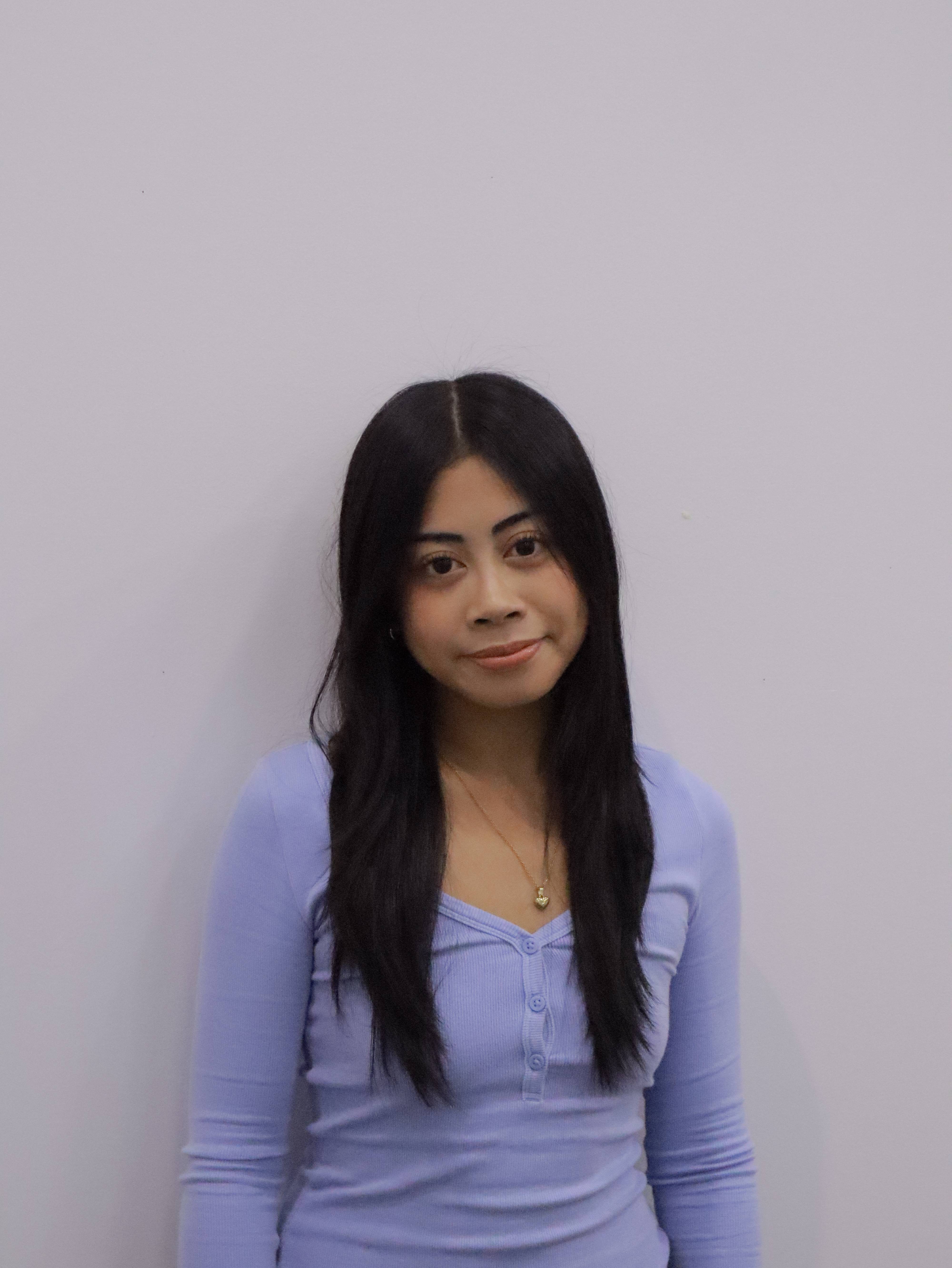 Woman with long black hair wearing a lavender buttoned shirt and gold heart necklace against a light gray background.