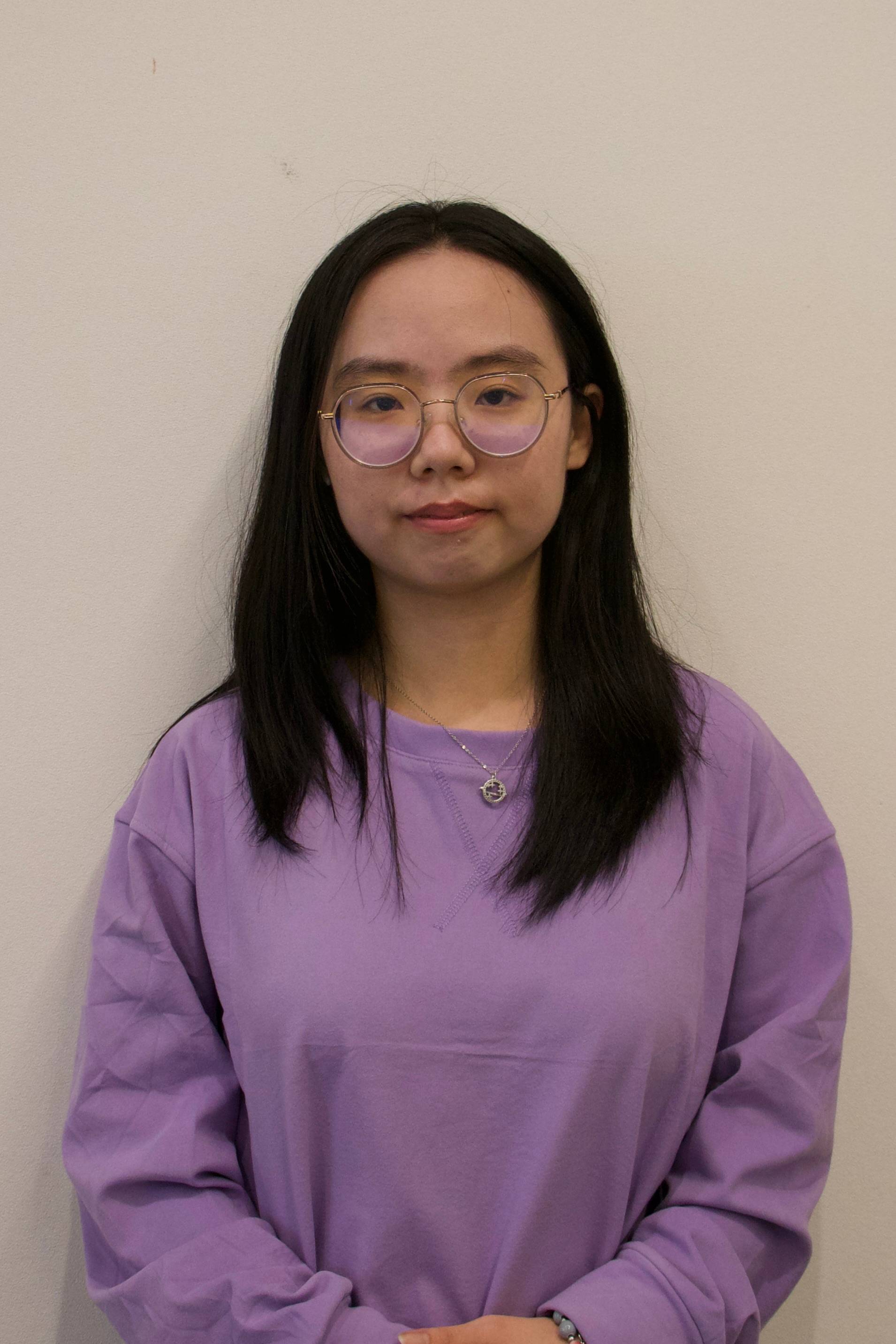 Young woman with long black hair wearing round glasses, a purple sweatshirt, a necklace, and a bracelet, standing against a plain wall.