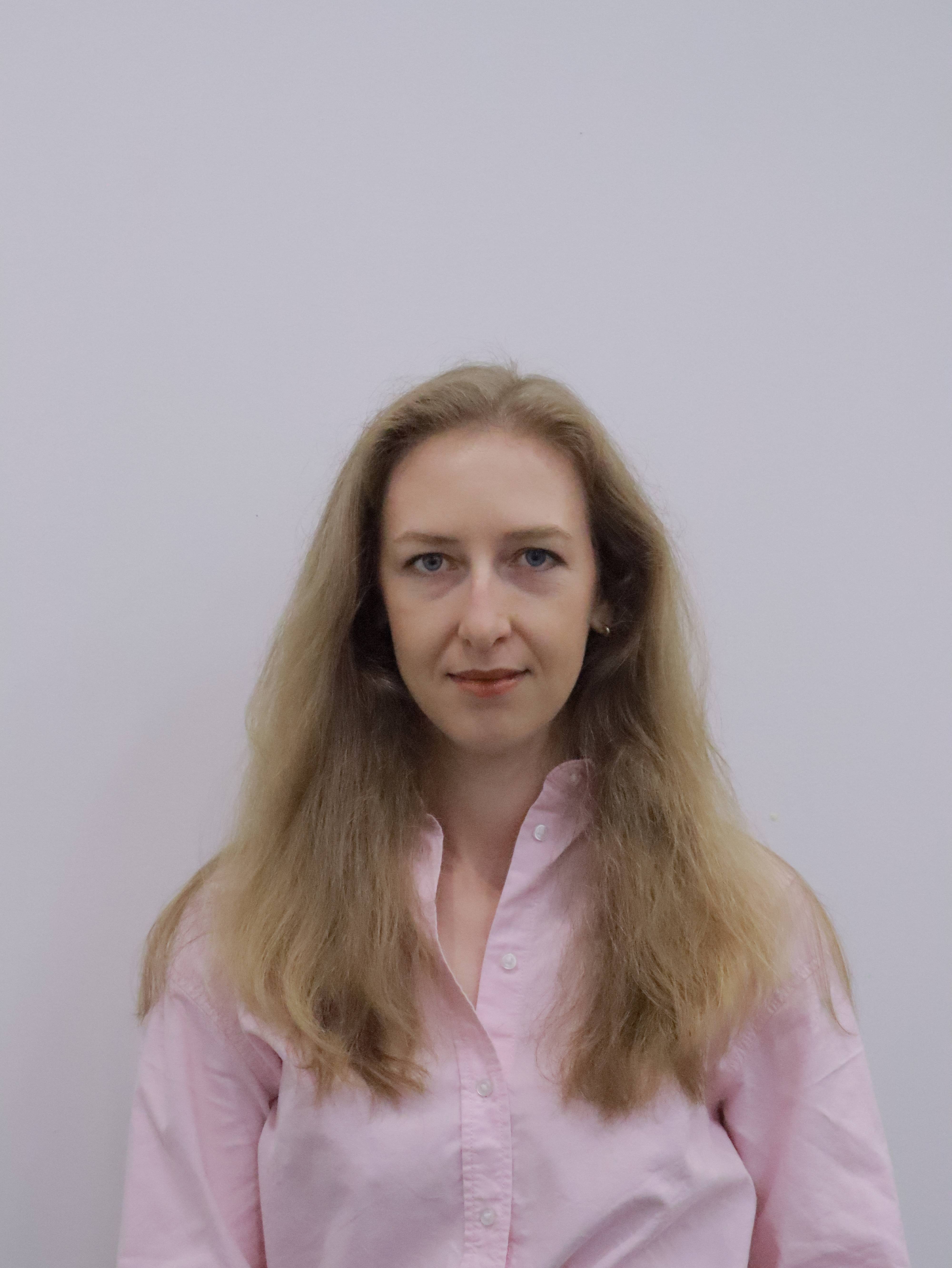 Woman with long light brown hair wearing a light pink button-up shirt against a plain light background.