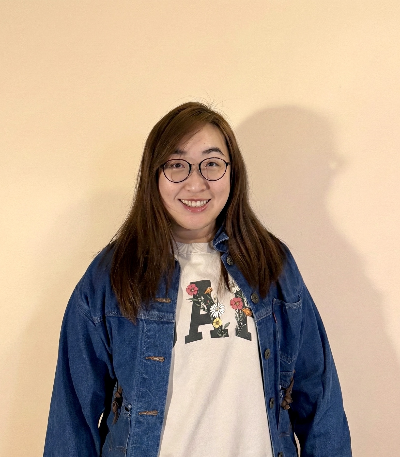 Smiling young woman with long dark hair wearing a denim jacket and light gray shirt against a plain beige background.