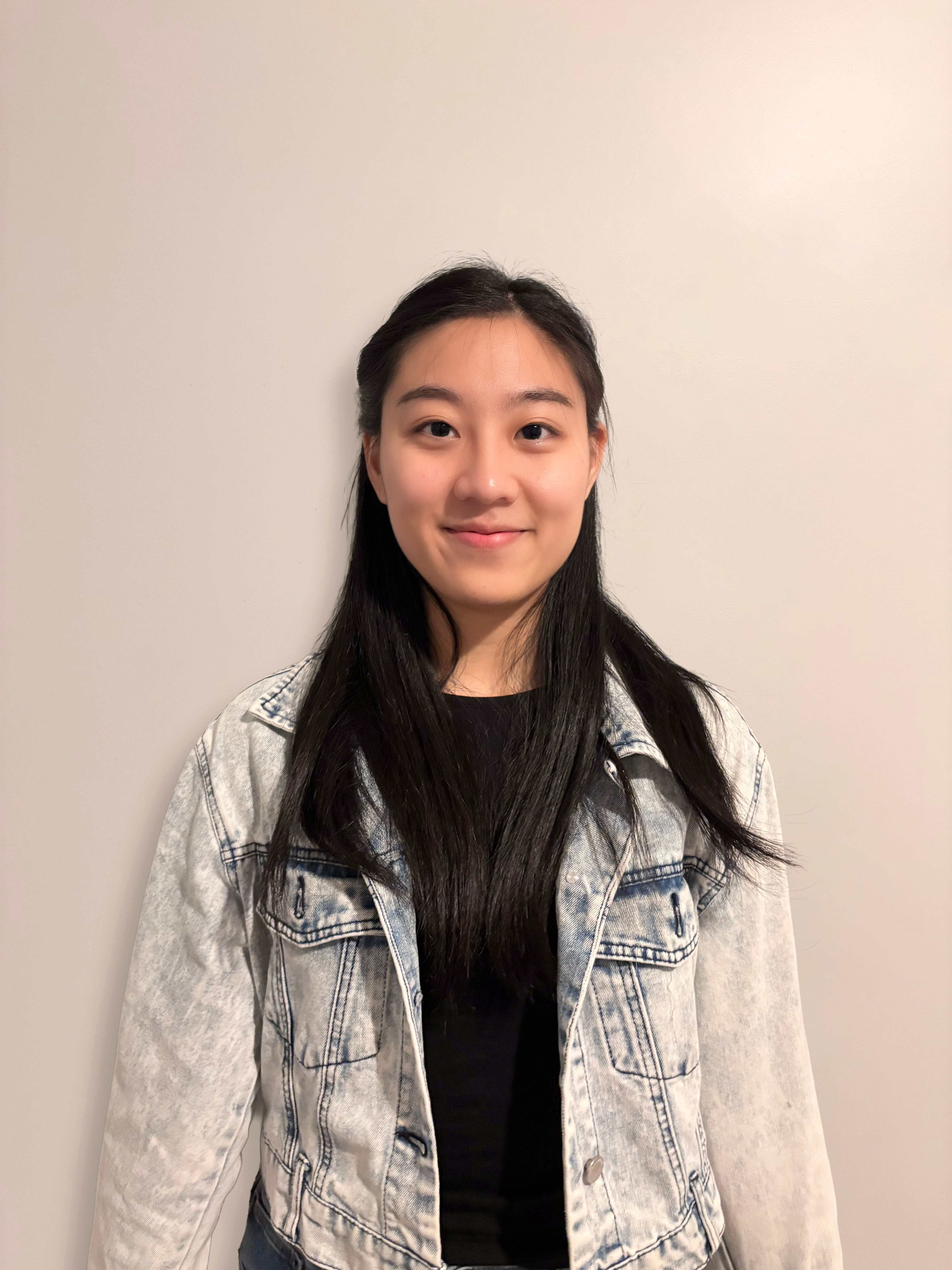 Smiling young woman with long dark hair wearing a denim jacket and light gray shirt against a plain beige background.
