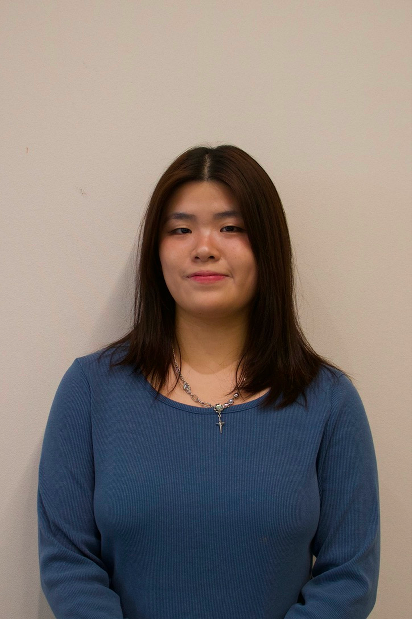 Smiling young woman with long dark hair wearing a denim jacket and light gray shirt against a plain beige background.