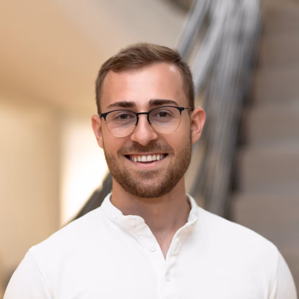 Smiling man with short brown hair, beard, and glasses wearing a white shirt indoors.