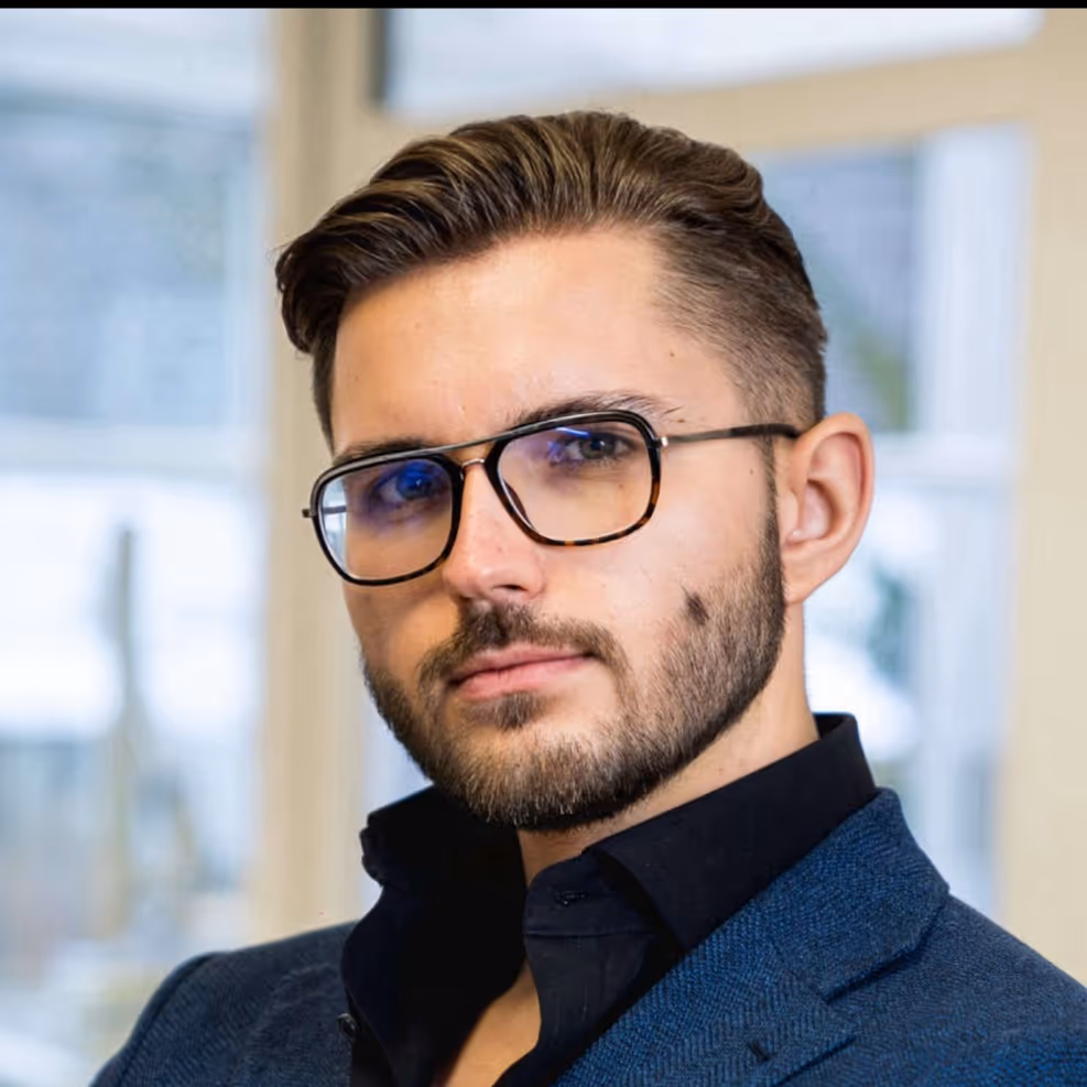 Man with short styled hair, beard, and glasses wearing a dark shirt and blue blazer indoors.