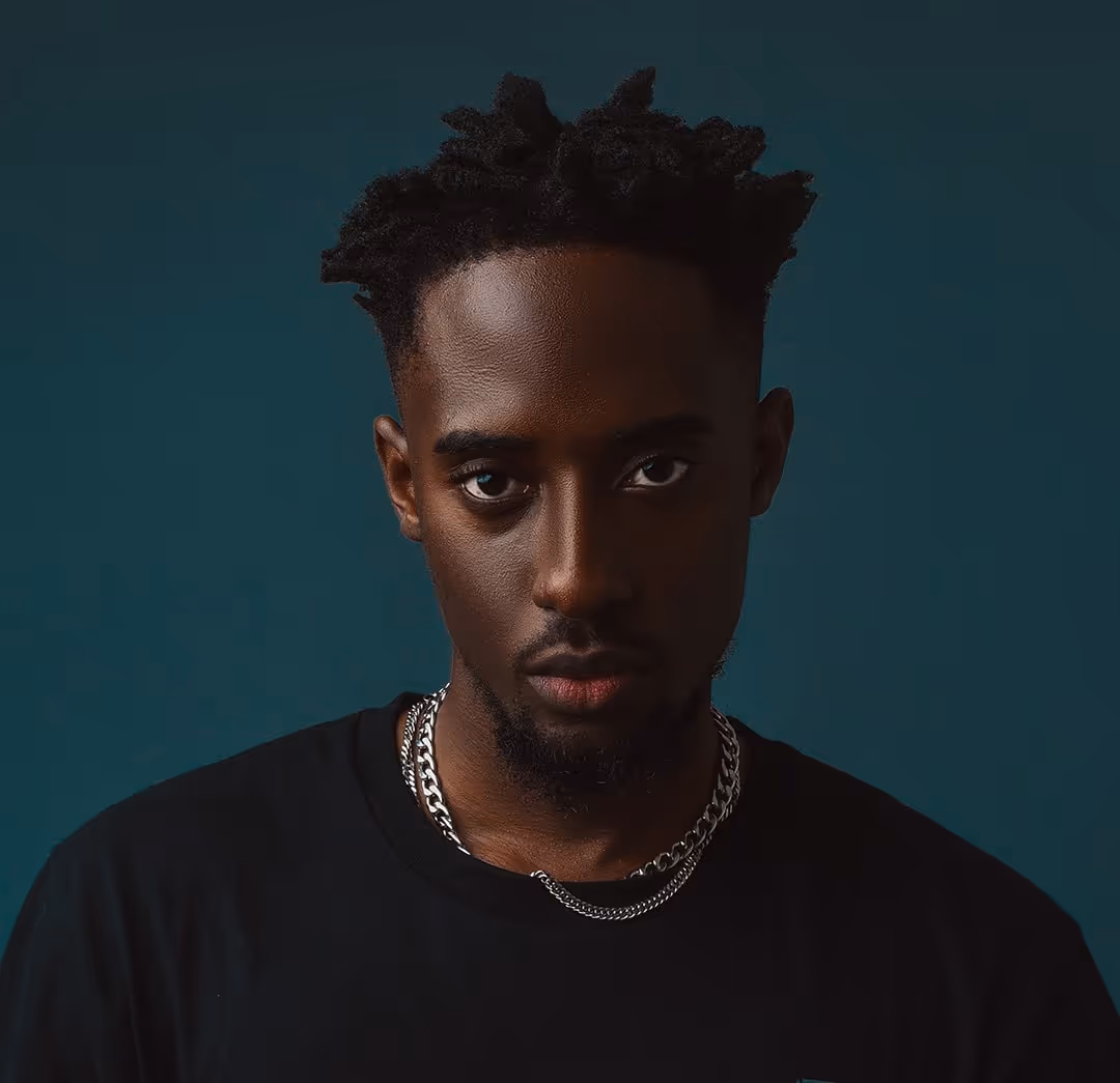Portrait of a young man with short twisted hair, a goatee, wearing a black shirt and silver chains against a dark teal background.