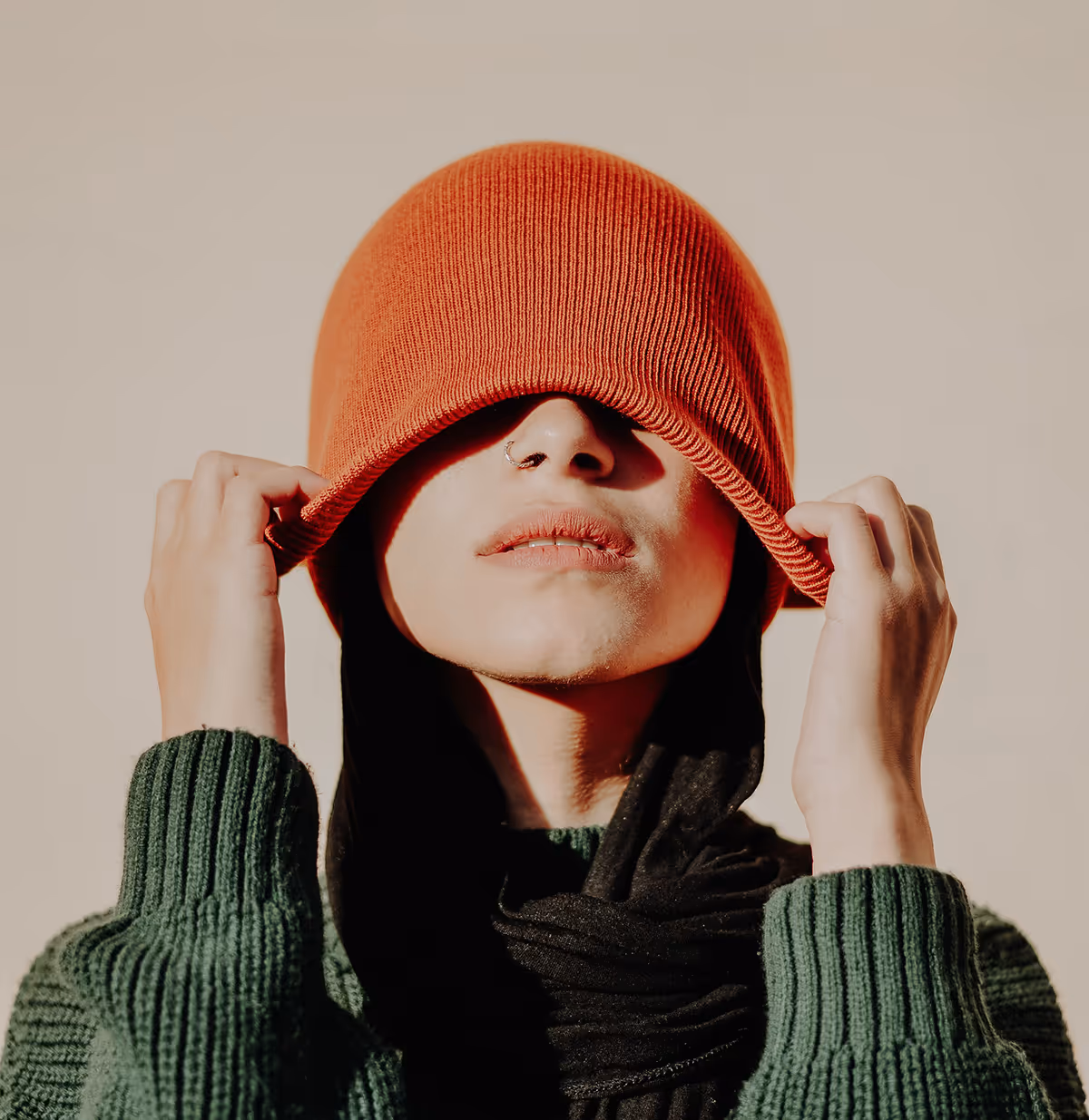 Person wearing an orange knit beanie pulled down over their eyes, a green sweater, and a black scarf.