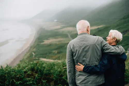 An older couple with their arms around one another looking over a foggy bay.