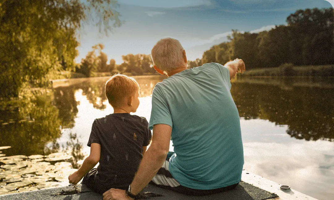 a child and his grandpa sitting next to a pond at sunset looking into the distance