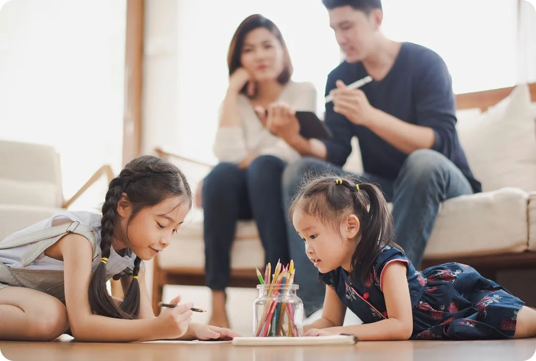 two children playing with colored pencils on the ground while two adults have a serious conversation in the background
