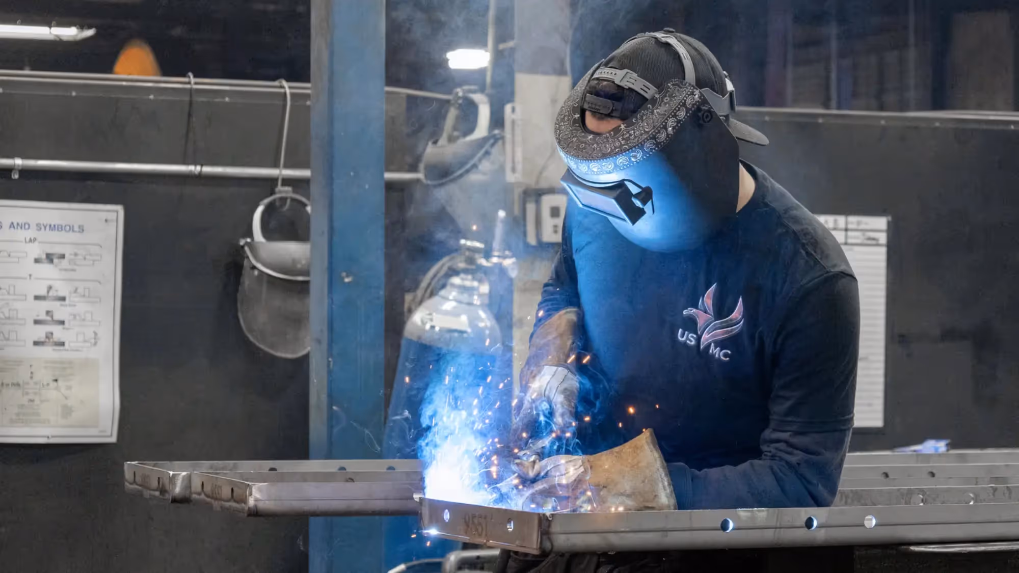 Worker in a welding helmet and gloves welds a metal frame at a workbench, with blue sparks and smoke in an industrial shop.