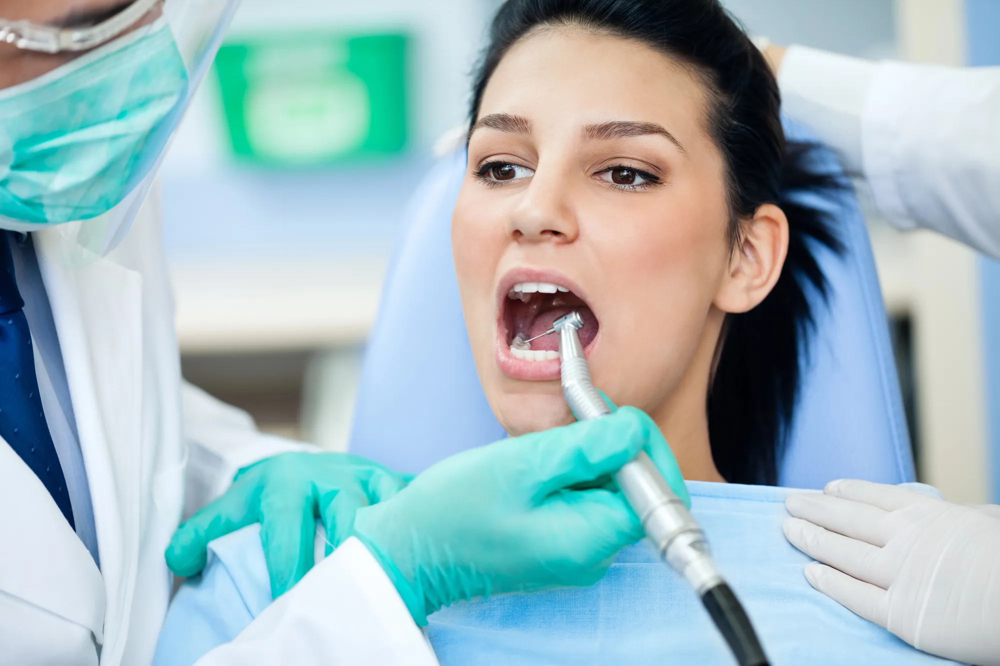 Dentist wearing gloves using a dental drill inside a young woman’s open mouth during a dental checkup.