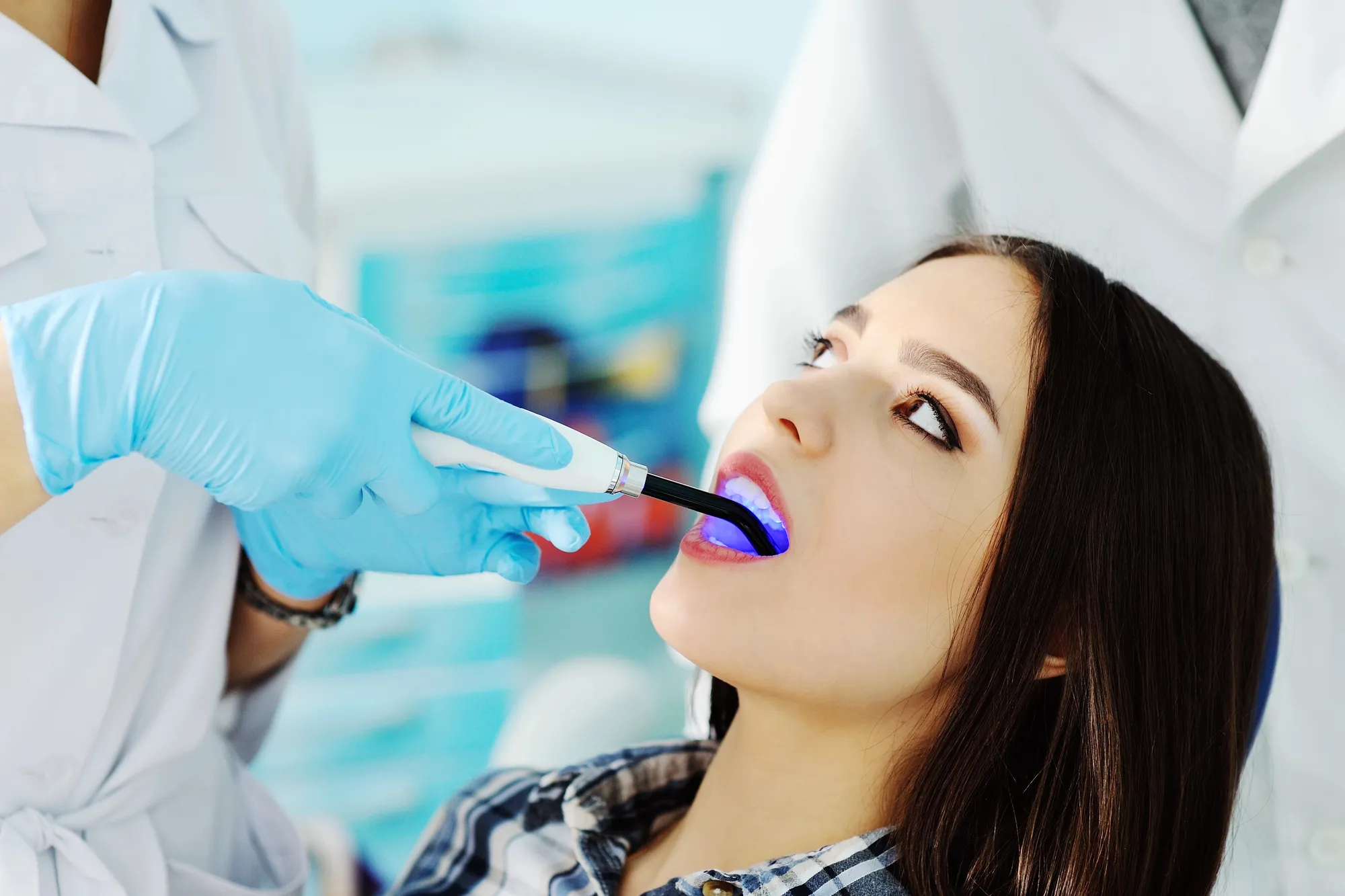 Dentist wearing blue gloves using a curing light inside a young woman's mouth during a dental procedure.