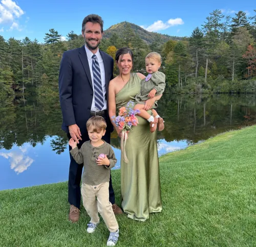 Family of four dressed formally standing on grass by a lake with trees and a mountain in the background.
