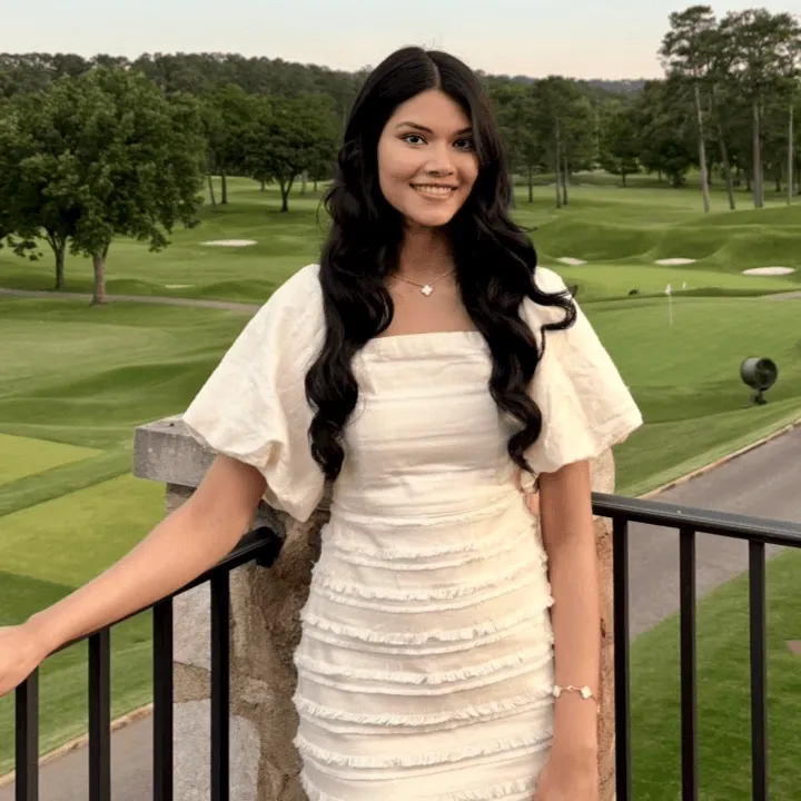 Woman with long dark hair wearing a white dress, smiling outdoors at a golf course in the evening.