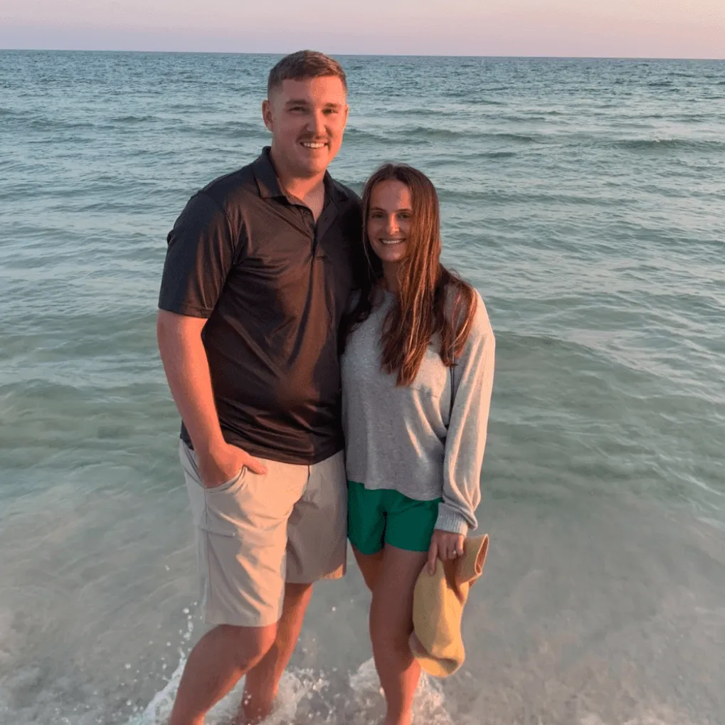 Smiling man in black shirt and woman in gray sweater with hat standing in ocean water at sunset.