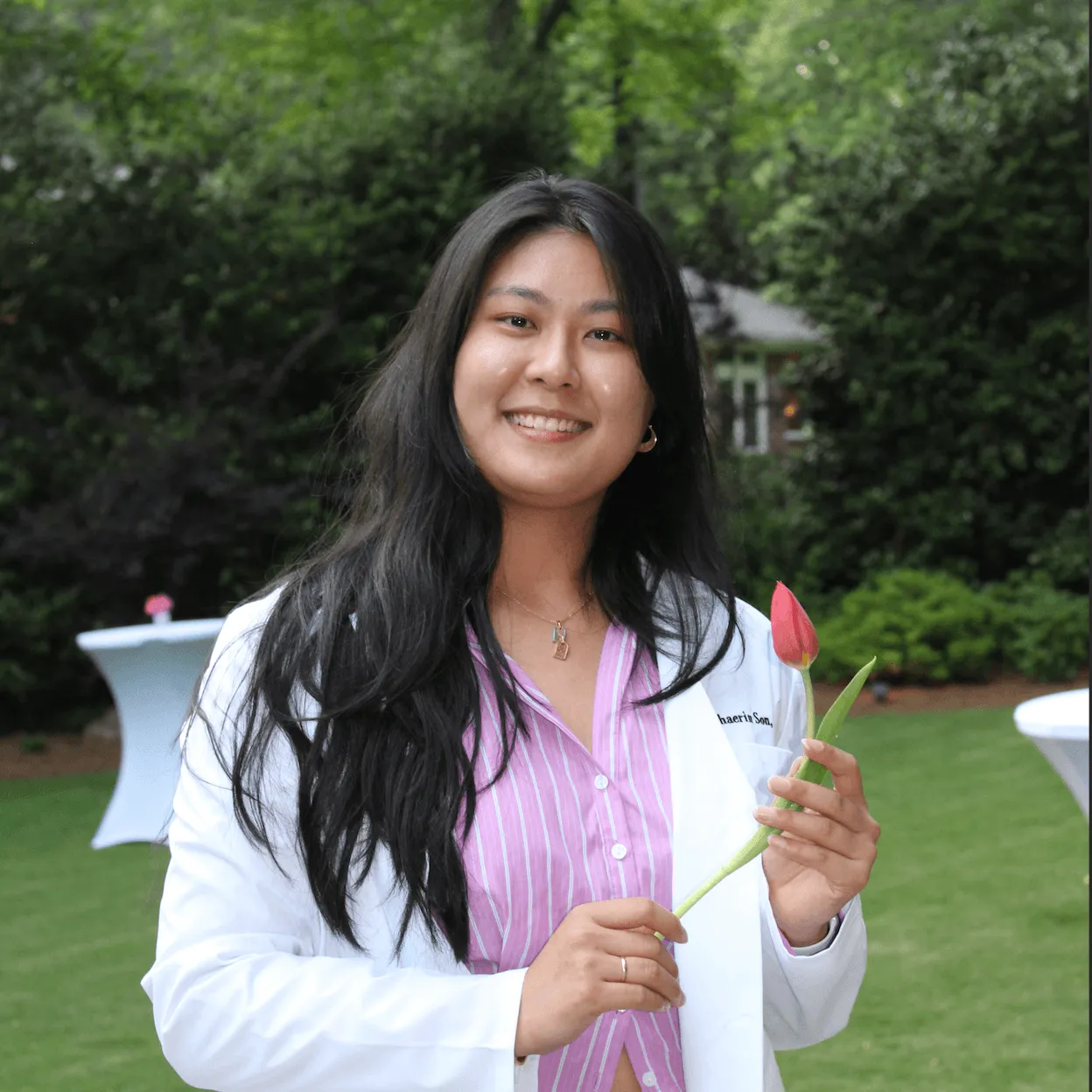 Smiling woman in white coat holding a red tulip in a green outdoor setting.
