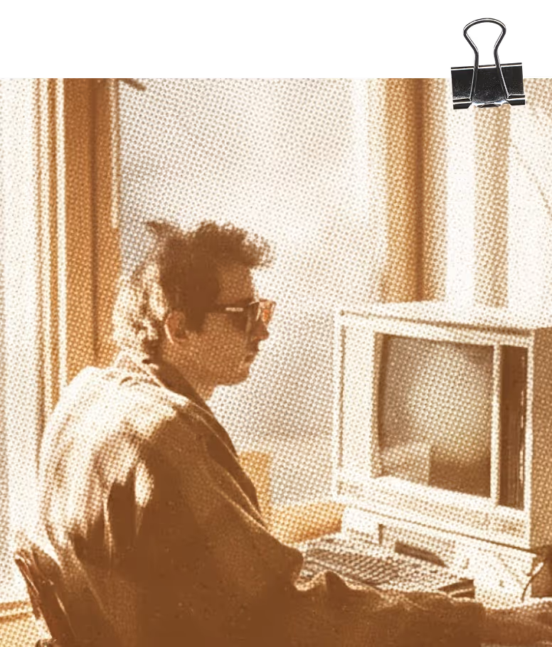 Person wearing sunglasses sitting at a desk using an old computer with a CRT monitor in a softly lit room.