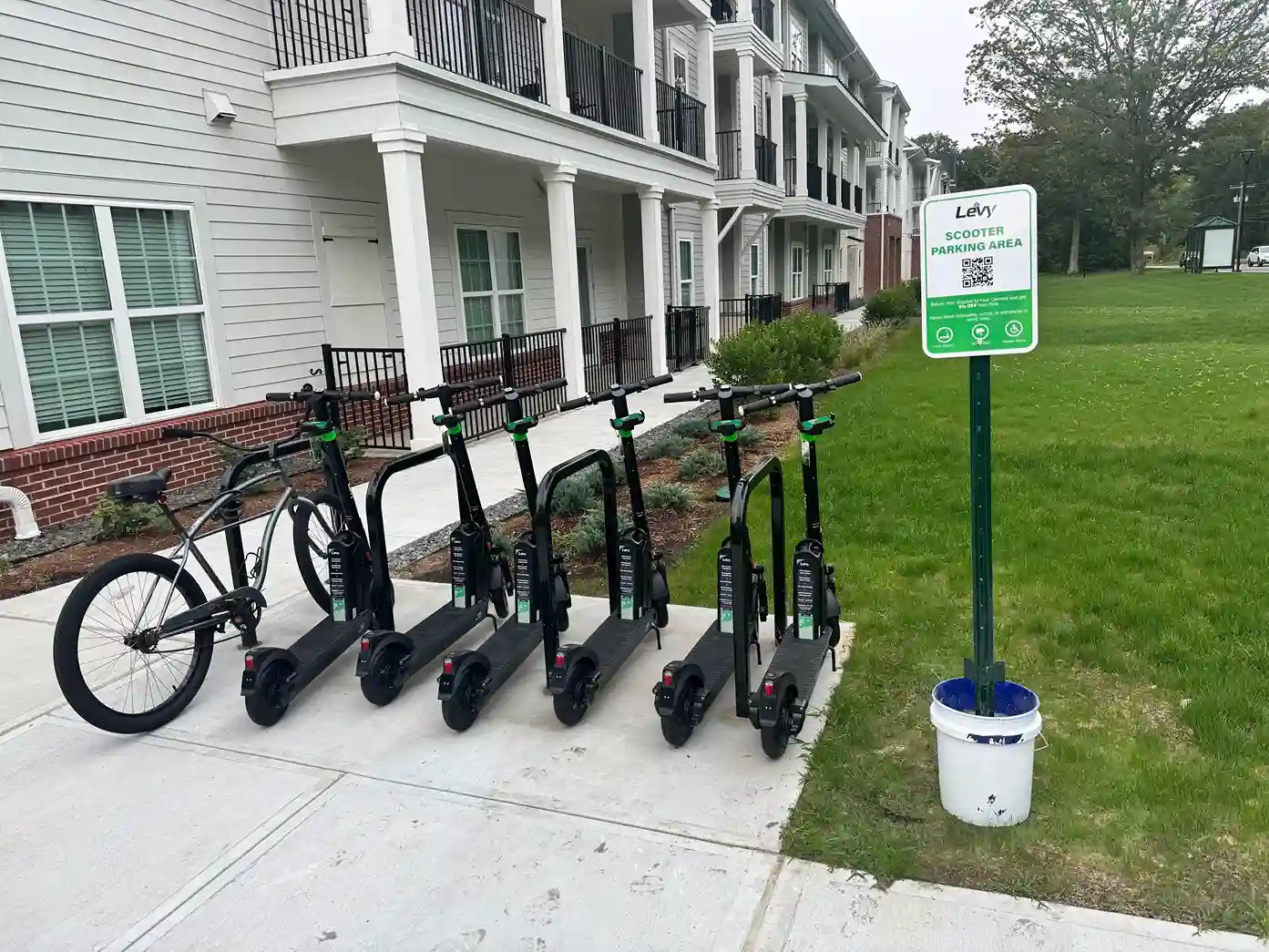 Five black electric scooters and one bicycle parked at a scooter parking area outside a residential building.