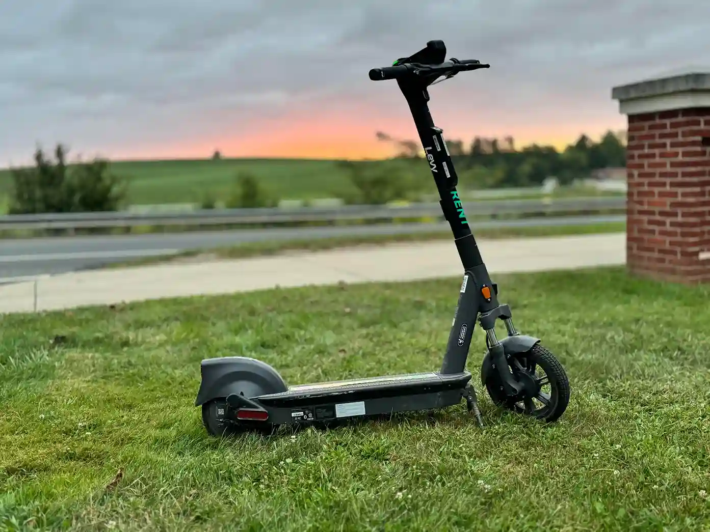 Black electric scooter parked on green grass near a sidewalk with a sunset and rural landscape in the background.