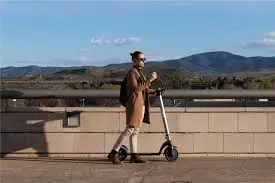 Man in brown jacket riding an electric scooter along a paved path with mountains in the background.