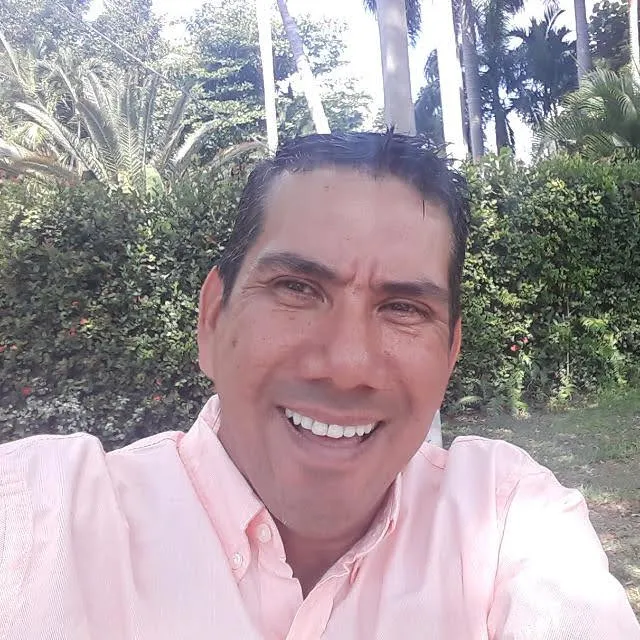 Smiling man in a light pink button-up shirt outdoors with green bushes and tall trees in the background.