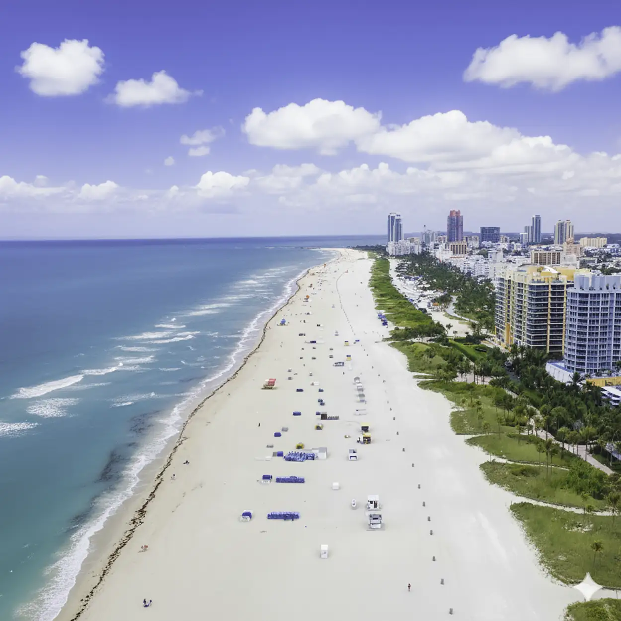 Aerial view of a long sandy beach with blue ocean waves on the left and high-rise buildings along the right side under a partly cloudy blue sky.