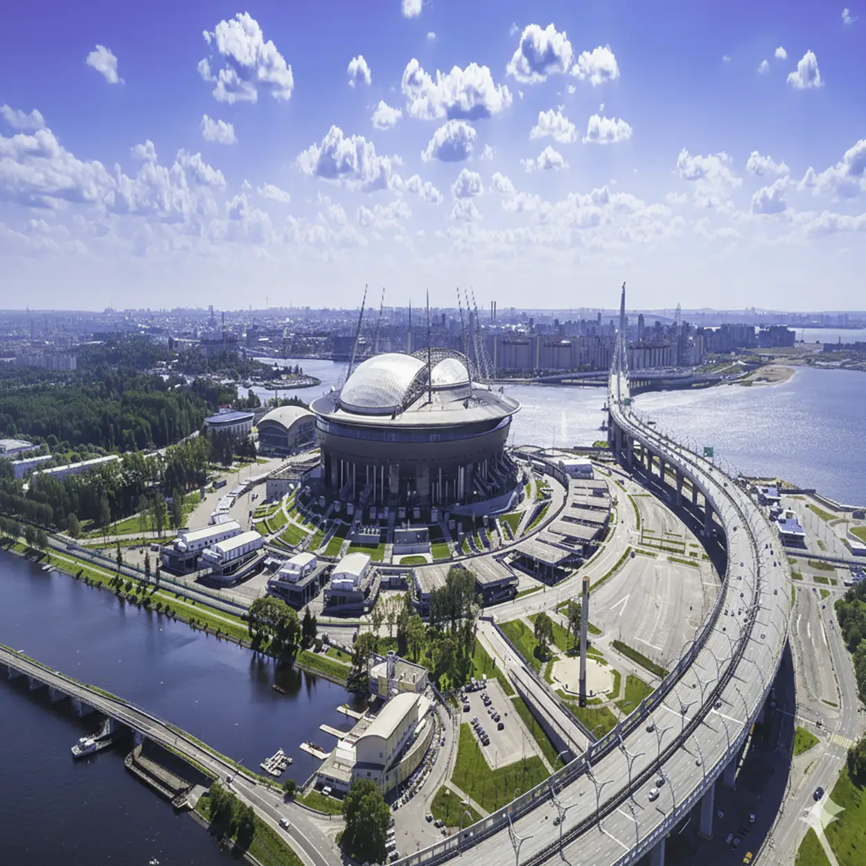Aerial view of a modern circular stadium with a partially open dome, surrounded by roads and bridges over water under a blue sky with scattered clouds.