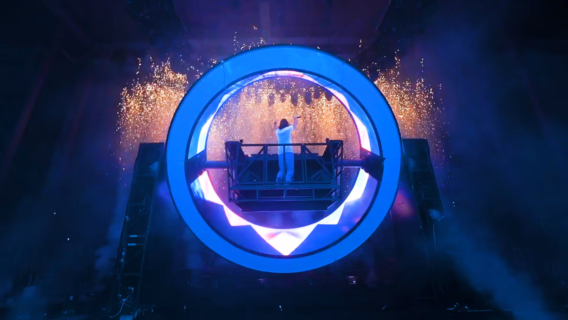 Performer in white outfit standing on a raised platform inside a large illuminated circular structure with fireworks in the background.