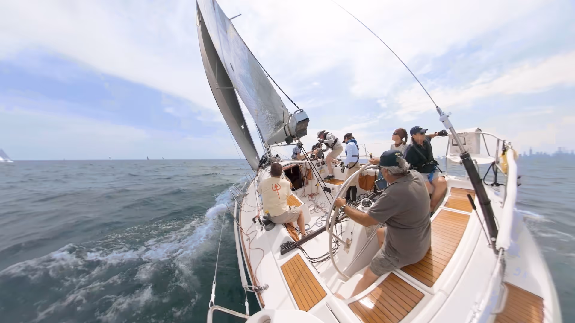 Crew of six people sailing on a white sailboat with brown wooden deck panels on a calm sea under a partly cloudy sky.