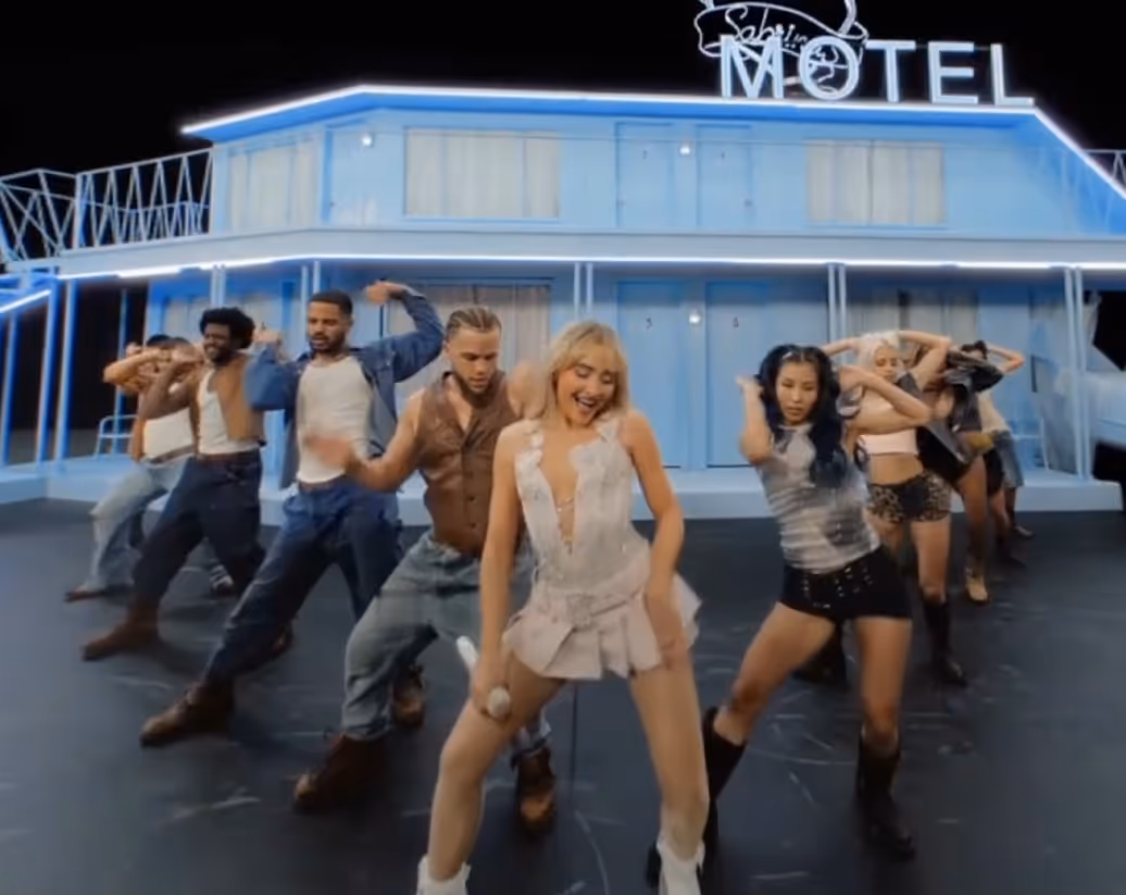 Group of dancers performing energetically in front of a blue neon-lit motel set.