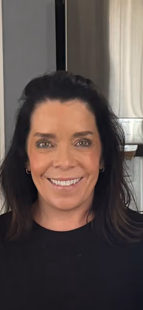 Smiling woman with dark hair and hoop earrings wearing a black top, indoors with curtains in background.