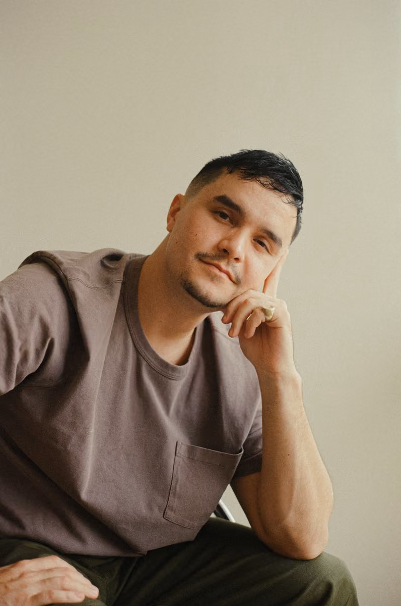 Man with short dark hair and goatee, wearing a brown shirt, resting his head on his hand.