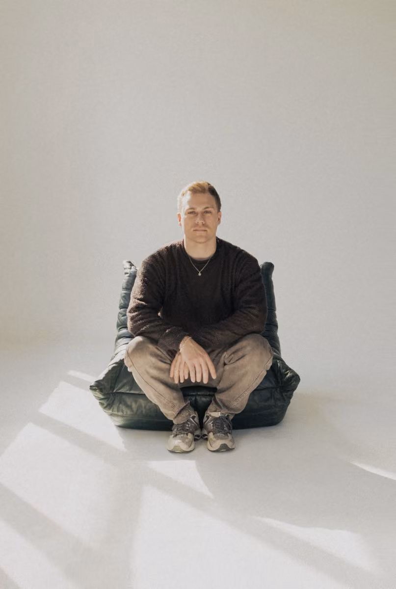 Man wearing brown sweater and beige pants sitting cross-legged on a dark cushion in bright room.