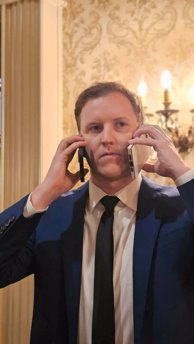 Man in a suit holding two smartphones to his ears in an ornate room with wallpaper and a chandelier.
