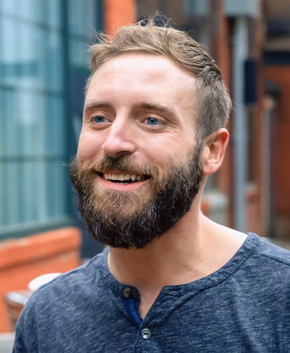 Smiling man with a beard and short hair wearing a blue shirt in an outdoor urban setting.