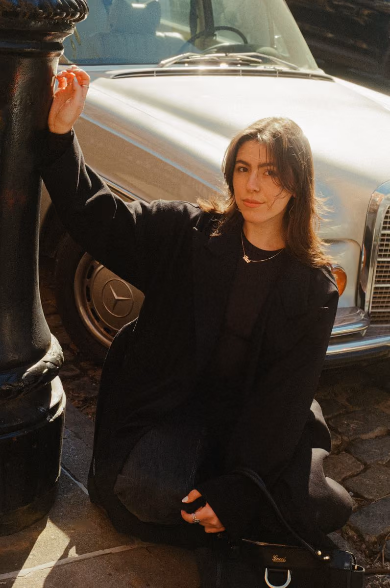 Woman in black coat sitting on pavement leaning on a lamppost with vintage Mercedes car behind.