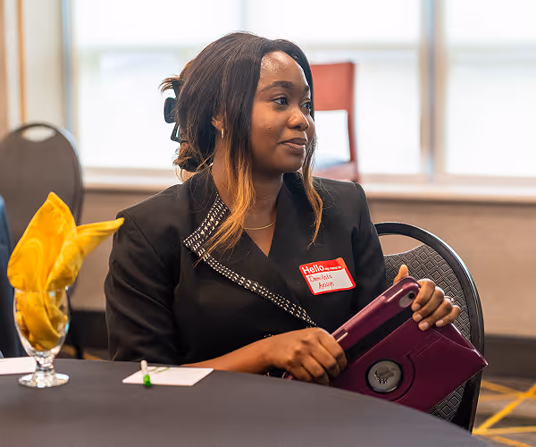 Woman with ombre hair holding a purple tablet, seated at a table with a yellow folded napkin and papers.