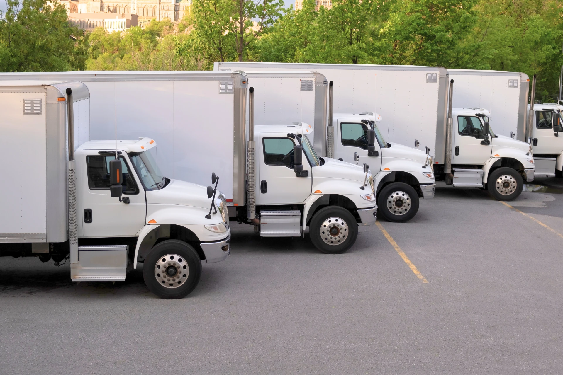 Four white box trucks parked side by side in a row in a paved lot, with green trees and buildings visible in the background