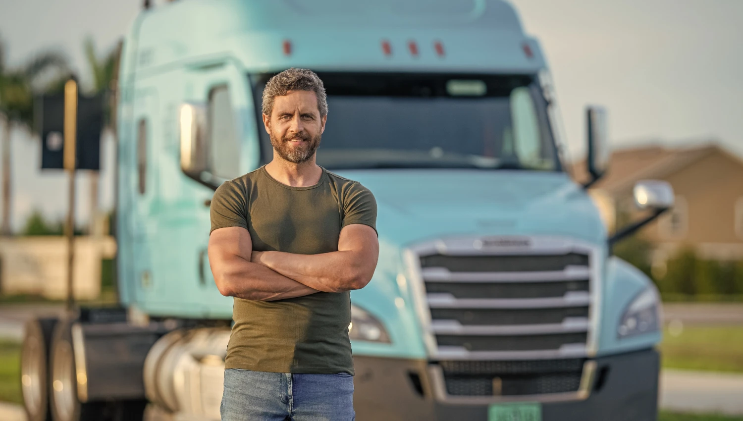 A trucker standing confidently with his arms crossed in front of a large light-blue semi-truck parked outdoors
