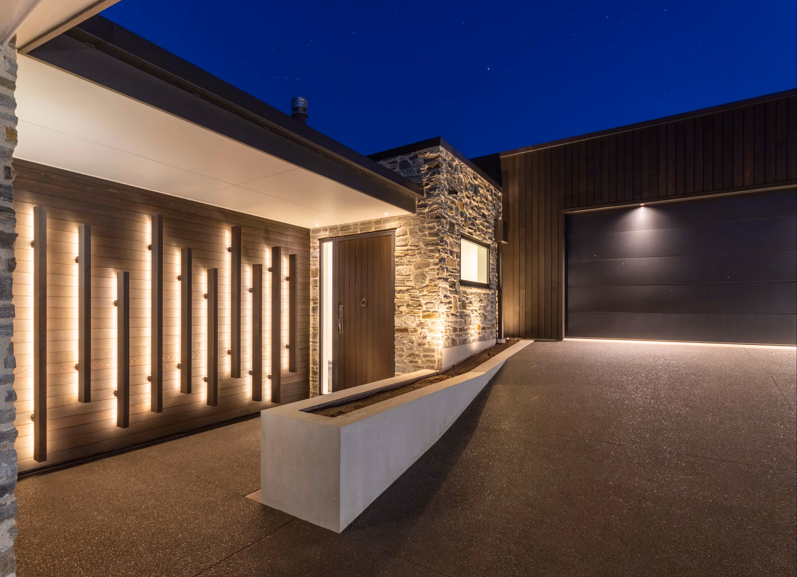 Modern house entrance at night with illuminated vertical wooden slats on the left wall, a brown door, stone siding, and a large dark garage door on the right.