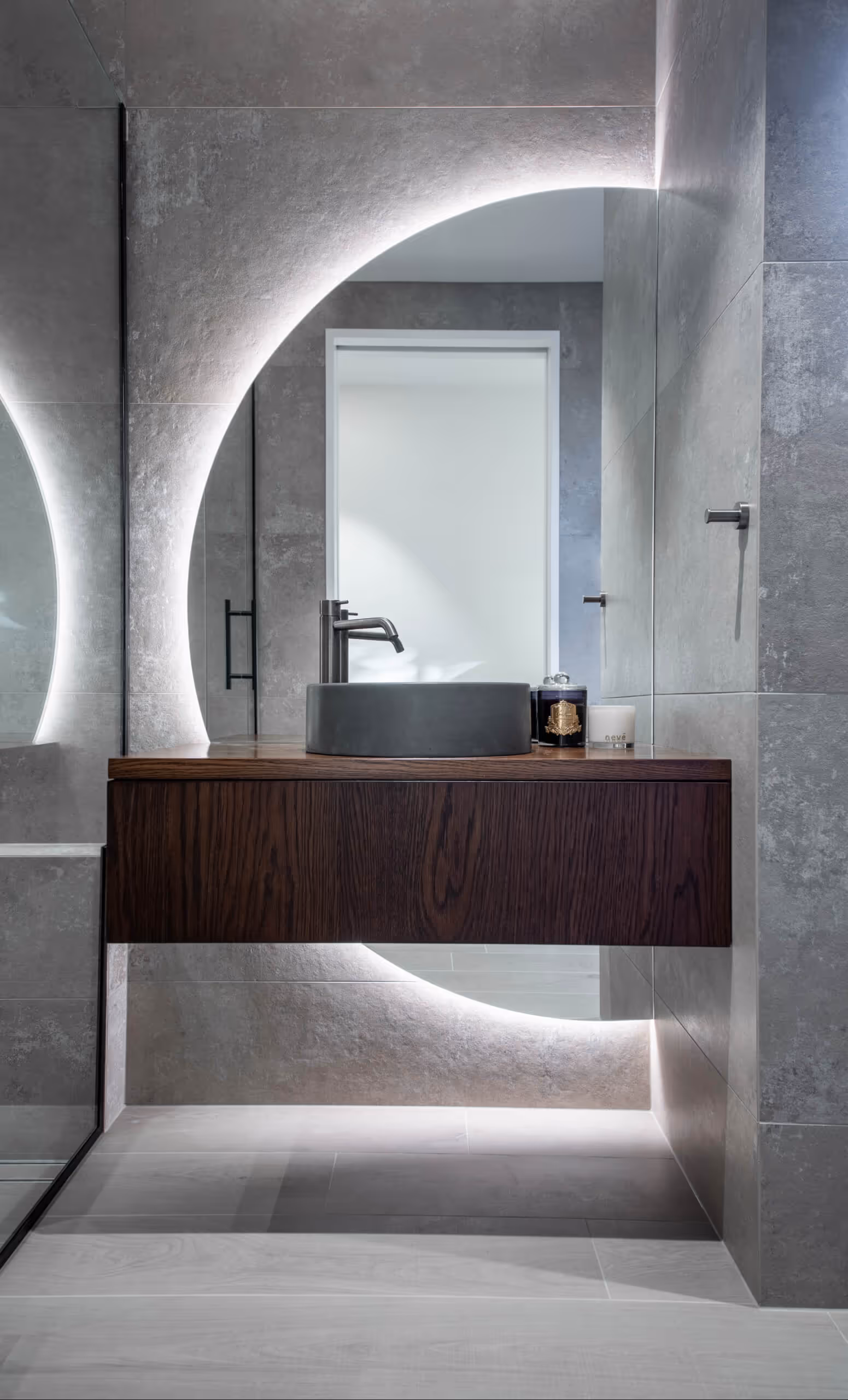 Modern bathroom with a round black sink on a floating wooden vanity and a large circular backlit mirror.